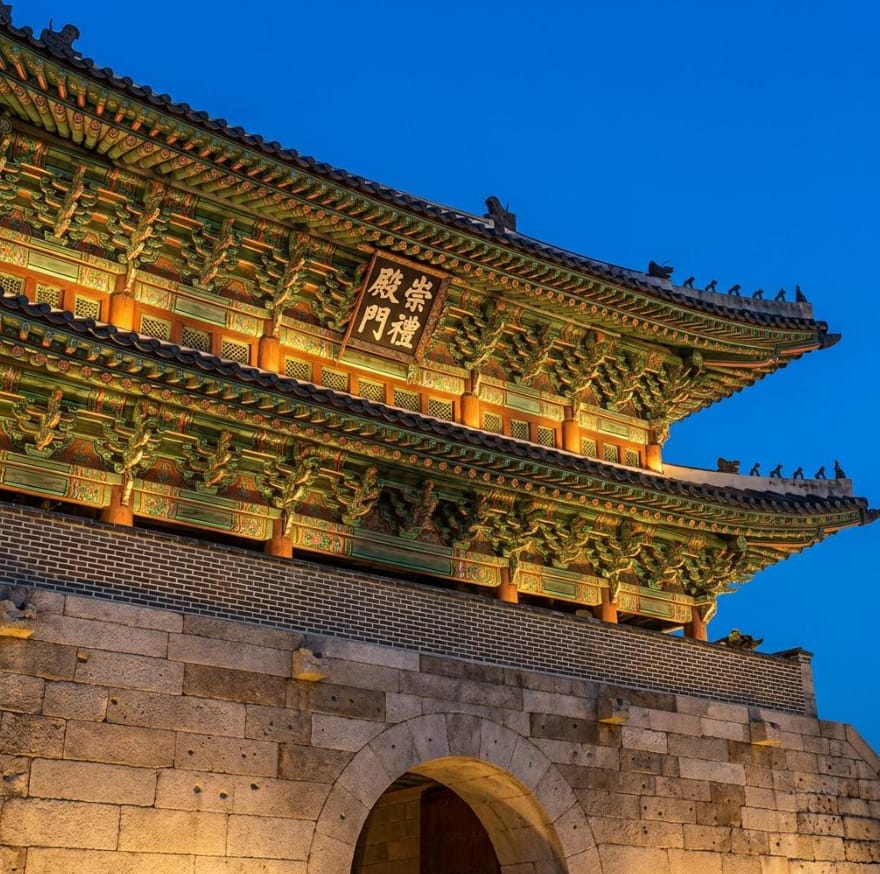 A dramatic night scene of Sungnyemun Gate engulfed in flames, with smoke billowing into the dark sky and firefighters helpless against the inferno.